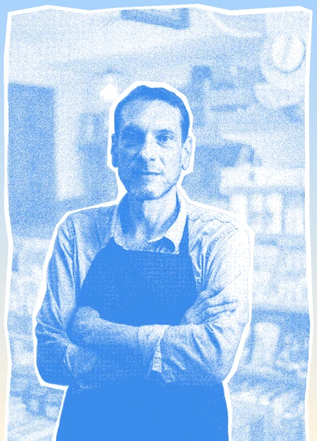 A shop owner in an apron standing with arms crossed in his store